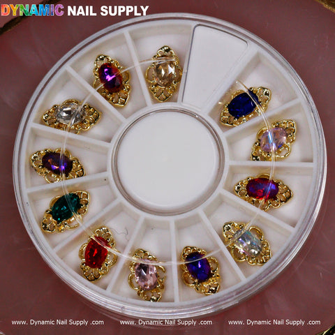The 12 pcs Luxury Nail Charms by Dynamic Nail Supply are arranged on a circular wheel, showcasing vibrant, gem-like decorations nestled in intricate gold settings. Each section contains a different colored charm—red, blue, pink, and clear—that is ideal for those wanting to explore dynamic nail design options. The setup is beautifully displayed against a soft pink background.