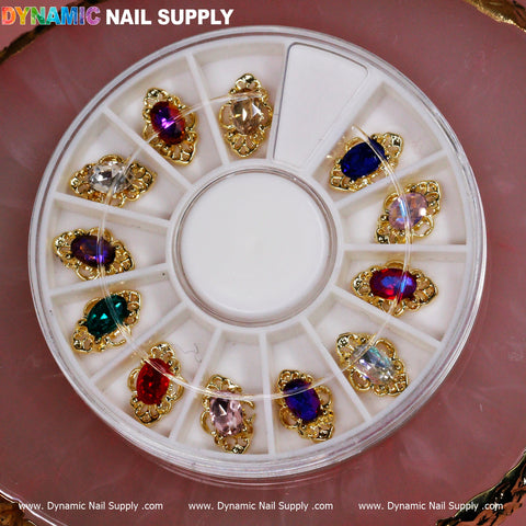 A circular plastic wheel from Dynamic Nail Supply holds luxurious nail charms in 12 compartments, each filled with jewel-like beads in a variety of colors, including red, purple, and clear, all set in gold-colored frames.