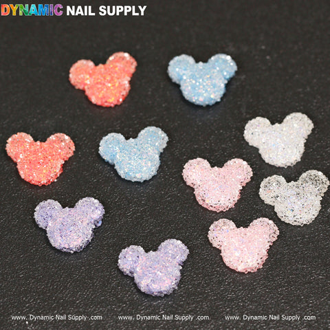 Scattered on a black surface are vibrant and glittery nail art decorations, similar to the 10 pcs Gummy / Sugar Candy Mickey Mouse Design Nail Charms from Dynamic Nail Supply. These delightful pieces come in shades of pink, blue, purple, and peach, each featuring a sparkly and textured appearance.