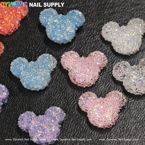 Dynamic Nail Supply's 10 pcs Gummy / Sugar Candy Mlckey Mouse Design Nail Charms are showcased on a textured black surface. Available in various colors like pink, blue, and clear, each charm sparkles with iridescent glitter for a delightful touch to your nails.