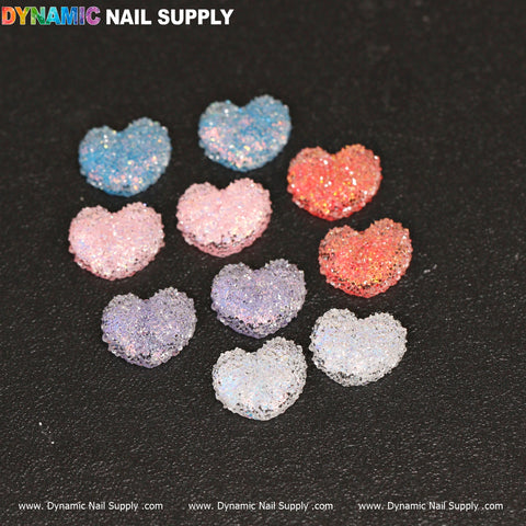 A set of ten glittery heart-shaped nail charms named "10 pcs Gummy / Sugar Candy Heart Shape Design Nail Charms" by Dynamic Nail Supply, available in a range of colors such as blue, pink, and silver. These charms are displayed on a black background, beautifully reflecting light, with the Dynamic Nail Supply logo positioned in the top left corner.