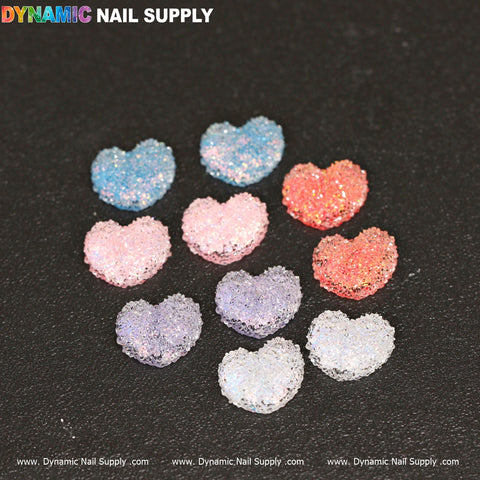 A collection of Dynamic Nail Supply's 10 pcs Gummy / Sugar Candy Heart Shape Design Nail Charms is displayed on a black background. These heart-shaped charms are arranged in a grid and come in various colors, including blue, pink, red, and purple, each featuring a glittery finish. The vibrant logo of Dynamic Nail Supply is prominently visible at the top.