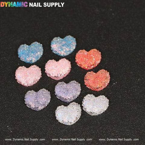 Ten vibrant heart-shaped nail charms from the 10 pcs Gummy / Sugar Candy Heart Shape Design Nail Charms set glisten against a black background. Arranged in a triangular formation, they display shades of blue, pink, red, purple, and white. The top edge of the image is adorned with the brand name Dynamic Nail Supply.