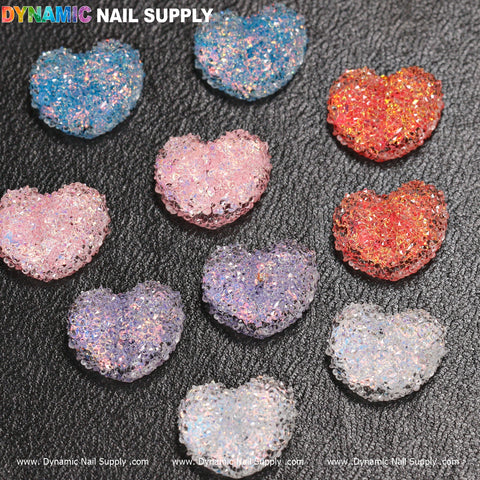 Ten gummy or sugar candy heart-shaped design nail charms from Dynamic Nail Supply, available in vibrant colors such as blue, pink, and purple, resemble dazzling candy-like nail decorations. Arranged neatly in three rows on a textured black background, they add sparkle and charm with "Dynamic Nail Supply" prominently displayed in the top left corner.
