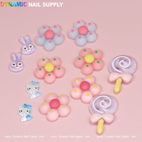 Against a pink background, vibrant nail art decorations stand out, featuring kawaii charms such as pastel flowers, adorable animal faces with bows, and lollipop designs from the 12 pcs Kawaii Candy Cartoon Mixed Shape Design Nail Charms collection. The brand Dynamic Nail Supply is prominently displayed at the top in rainbow letters, with their website repeated below.