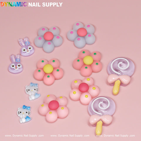 A collection of 12 pcs Kawaii Candy Cartoon Mixed Shape Design Nail Charms displayed against a light pink background, showcasing adorable designs such as bunnies, cats, flowers, and lollipops. The pastel colors contribute to a playful vibe. Bunnies and cats are adorned with tiny bows, while the lollipops feature pink bows and yellow sticks. The Dynamic Nail Supply branding text is displayed at the top.