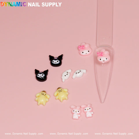 The 10 pcs Cute Cartoon Character Design Nail Charms from Dynamic Nail Supply embellish a light pink background showcasing black cats with white faces, yellow ducklings, and pink rabbits with bows. Some charms are displayed on clear plastic nail samples, while the bottom edge features Dynamic Nail Supply logos for a branded finish.