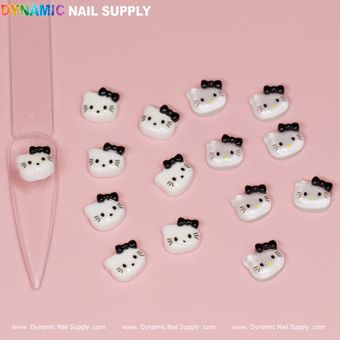 On a pink surface, there are 14 small, white nail charms from the Dynamic Nail Supply collection, each featuring a Hello Kitty design with a black bow on one ear. These cats display varied expressions using black dots and lines for eyes, noses, and whiskers. A transparent nail tip by Dynamic Nail Supply is positioned to the left.