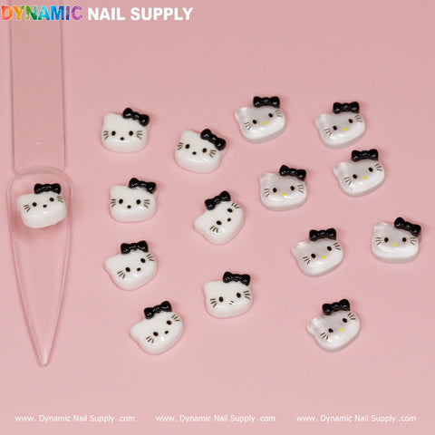 A collection of 15 small, white Hello Kitty design nail charms featuring black bows on their ears, artfully arranged on a pink background. One charm is showcased on a clear nail tip. The brand name "Dynamic Nail Supply" appears in vibrant colors at the top and is repeated in white at the bottom.