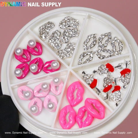 A close-up of a round container featuring six compartments filled with vibrant nail decorations. The sections include eye-catching nail charms such as silver rhinestone lips, pink lips, 3D Lips Nail Charms, pink bows with pearls, and small red cherries embellished with silver rhinestones. The background is a pink hue with the Dynamic Nail Supply brand displayed at the top.