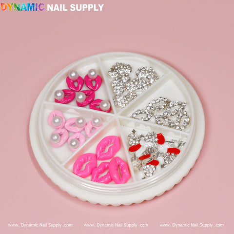 On a pink background rests a circular nail art palette featuring six compartments containing decorative nail charms, such as pink gems, pearls, 3D Lips Nail Charms, and silver rhinestones. The brand name "Dynamic Nail Supply" is prominently displayed on the packaging.