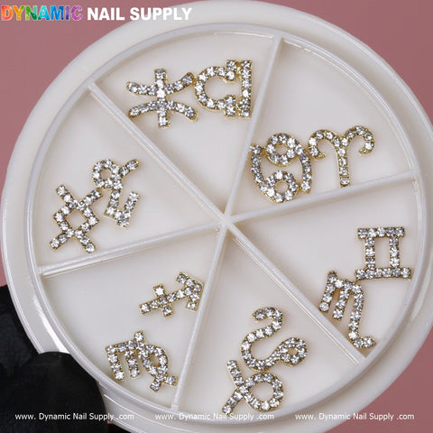 A circular container features eight compartments, each containing Gold Zodiac Sign Nail Charms with rhinestone detailing from the Dynamic Nail Supply collection. Included in this set are charms representing Virgo, Aquarius, Pisces, Aries, Gemini, Taurus, Sagittarius, and Capricorn against a soft pink backdrop.
