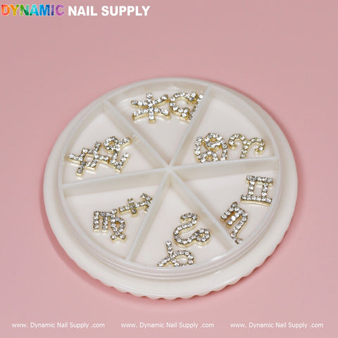 A round white nail art wheel, positioned on a pink surface, features the Set of 12 pcs Gold Zodiac Sign Nails Charm with Rhinestones, each neatly organized in its own compartment. The lid displays the brand name Dynamic Nail Supply.