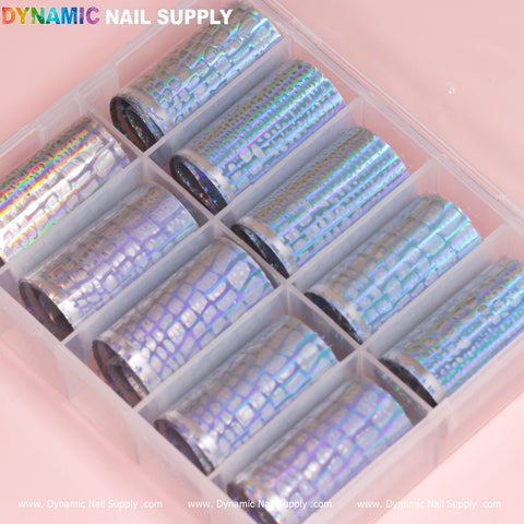 The Nail Art Design Foils - Animal Skin Holographic from Dynamic Nail Supply are enclosed in a transparent plastic box containing ten rolls of foils with a scale-like pattern. These holographic nail art foils exhibit an iridescent sheen, reflecting a spectrum of colors. The box is set against a soft pink backdrop, with "Dynamic Nail Supply" printed in vibrant letters above.