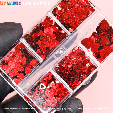 A person wearing black gloves holds a plastic case from Dynamic Nail Supply. The case features six compartments filled with the Red Metallic Valentine Style Mix-sizes Heart Shape Sequins Glitter, offering a variety of red heart-shaped decorations. Against a soft pink background, the container showcases various styles for creative manicures.