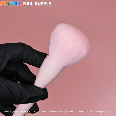 A hand wearing a black glove holds a fluffy Soft-pink Dust Brush (Cosmetic-grade Duster) against a solid pink background. The brush features a pale pink handle and soft bristles. The text Dynamic Nail Supply is displayed in colorful letters at the top left corner.