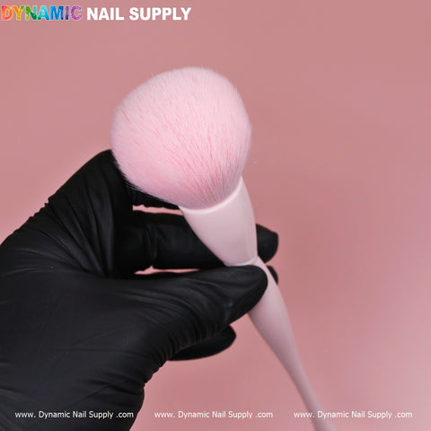 A hand in a black glove presents a Soft-pink Dust Brush featuring a white handle and fluffy pink bristles against a solid pink background. The text "Dynamic Nail Supply" appears in colorful letters in the top left corner, while the bottom showcases the URL www.DynamicNailSupply.com repeatedly, emphasizing its cosmetic-grade quality.