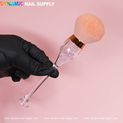 A gloved hand holds the Wine-cup style Dust Brush, a cosmetic-grade duster from Dynamic Nail Supply. This elegant accessory features a long, transparent handle filled with sparkling crystal boba and soft, fluffy bristles with a stunning metallic rose-gold accent at the base. The background is a soft pink, and the name "Dynamic Nail Supply" is displayed in the upper left corner.