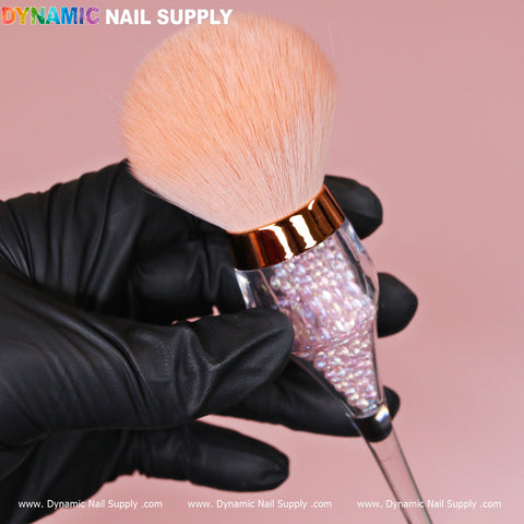 A hand in a black glove holds an ornate, cosmetic-grade Wine-cup style Dust Brush from Dynamic Nail Supply, featuring fluffy light peach bristles. Its transparent handle is beautifully filled with small, shiny Crystal Boba beads. The scene is set against a soft pink background, with the top left corner displaying the brand name Dynamic Nail Supply, highlighting this elegant rose-gold duster.