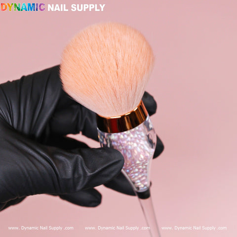 A gloved hand holds the Wine-cup style Dust Brush from Dynamic Nail Supply, featuring a large, fluffy light peach-colored bristle head. The iridescent rose-gold handle with crystal boba inside and a golden accent near the bristles resembles a dust brush. The background is soft pink, with "Dynamic Nail Supply" in colorful letters at the top.