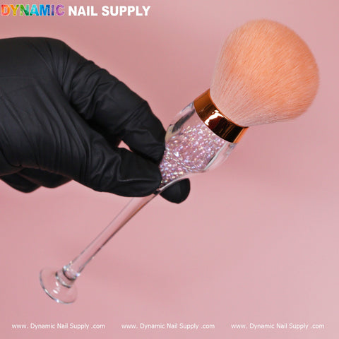 A hand clad in a black glove holds the Wine-cup style Dust Brush, a cosmetic-grade duster from Dynamic Nail Supply, featuring fluffy peach-colored bristles. Its transparent handle is adorned with sparkling glitter and complemented by a metallic rose gold ferrule. The scene is set against a solid pink background, with "Dynamic Nail Supply" prominently displayed in bold, colorful letters at the top left corner.