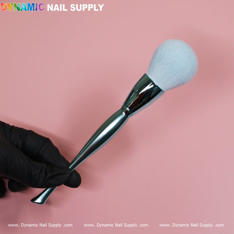 A hand in a black glove holds a Dynamic Nail Supply Sky Blue Dust Brush, featuring a metallic design and white bristle head, against a pink background. The text DYNAMIC NAIL SUPPLY appears at the top, with website URLs repeated below. This cosmetic-grade duster boasts an ergonomic, sleek design.