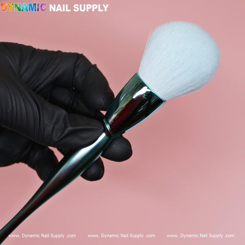 A gloved hand holds a sleek, metallic Sky Blue Dust Brush with a large, soft white bristle head against a pink background. At the top left corner are the words Dynamic Nail Supply, while the bottom repeats: www.DynamicNailSupply.com. Perfect for cosmetic-grade precision.