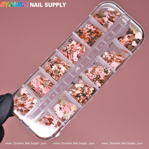 A hand holds a clear plastic case filled with compartments displaying the Pink Gold Mix-sizes Butterfly & Flower & Heart shape sequin glitter. The sparkling sequins feature small hearts, stars, and hexagons in pink and gold tones. Against a solid pink background, the Dynamic Nail Supply logo is prominently positioned at the top left corner.