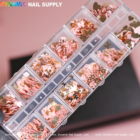 The clear plastic organizer case from Dynamic Nail Supply houses a collection of nail art decorations featuring gold butterfly, flower, and heart-shaped sequins and glitter. The box is designed with individual compartments arranged in two rows against a soft pink background. The product is known as the Pink Gold Mix-sizes Butterfly & Flower & Heart shape sequin glitter.