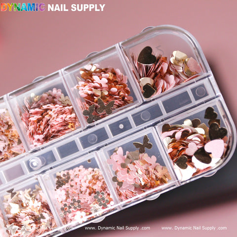 A clear plastic case houses an assortment of small metallic nail art decorations from Dynamic Nail Supply. The sparkling sequins are in various shapes such as butterflies, flowers, and hearts, and come in mix-sizes with a dazzling array of pink, gold, and silver shades set against a soft pink background.