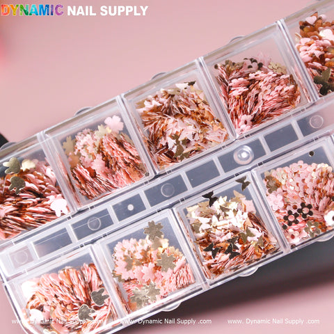 A plastic organizer case from Dynamic Nail Supply, named the Pink Gold Mix-sizes Butterfly & Flower & Heart shape sequin glitter, features multiple compartments filled with assorted nail art decorations. The contents boast metallic rose gold and gold shapes including stars, flowers, and leaves, all arranged alongside glimmering sequins. The case has a soft pink background with a partially transparent lid.