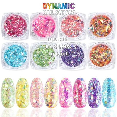 The image displays a set of eight holographic nail art glitter powders from Dynamic Nail Supply, each in clear containers. The vibrant colors include red, pink, orange, purple, green, blue, yellow, and multicolor. Below the containers are swatches illustrating how the powders appear when applied on nail shapes. The text reads "QH15060 SET 02 Holographic Nail Art Glitter Set Powder" by Dynamic Nail Supply with 1g of each color.

