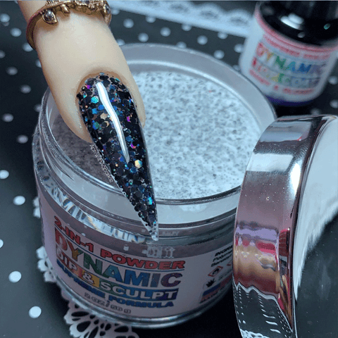A hand displaying long black tips adorned with holographic glitter carefully dips into an open jar of Black Holographic Glitter Nail Powder #322 by Dynamic Nail Supply. The jar is set against a sophisticated black and white polka dot backdrop. In the background, a bottle featuring the same design suggests endless opportunities for creative 3D nail art. A gold ring elegantly graces the hand.