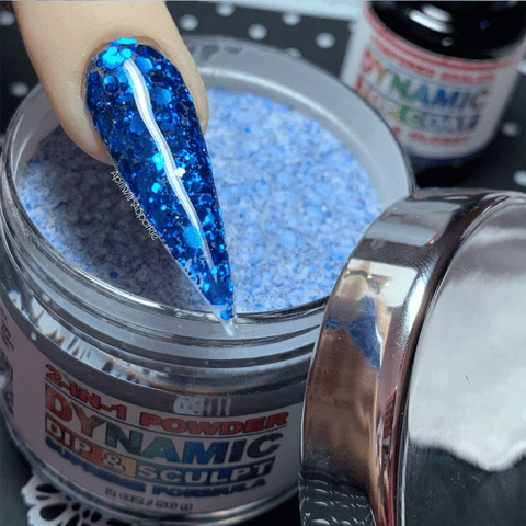 A hand with a long, pointed nail is gracefully dipped into a jar of Blue Holographic Glitter Nail Powder #372 by Dynamic Nail Supply. Coated in non-toxic shimmer, the nail stands out against the decorated surface. In the background, another jar from Dynamic Nail Supply awaits exploration for creating stunning nail designs.