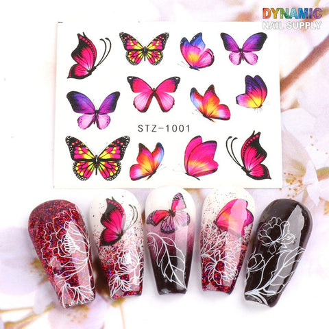 Discover the Dynamic Nail Supply Designer Nail Art Set, featuring a pack of vibrant butterfly water decals for 3D nail art decoration. This set includes stunning designs with pink, purple, and orange butterflies on five artificial nails that showcase intricate white floral and butterfly patterns over red, pink, and black gradients. The backdrop highlights a beautifully blurred floral design.
