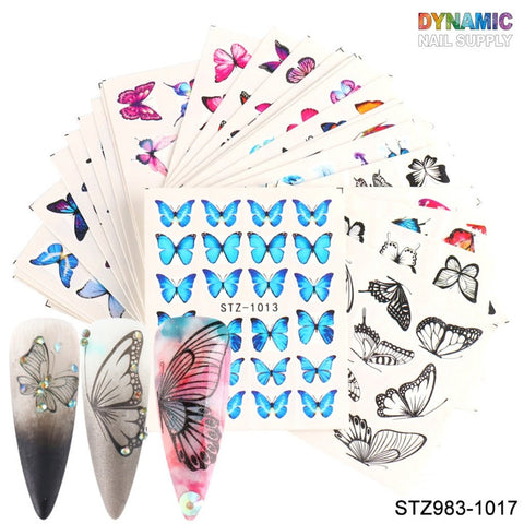 A collection of 24 sheets from Dynamic Nail Supply's Butterfly Water Decals features 3D designer nail art with vivid blue, pink, and multicolored patterns, elegantly displayed in a fanned presentation. At the bottom, three nail samples exhibit intricate butterfly designs on gradient backgrounds adorned with rhinestone accents.