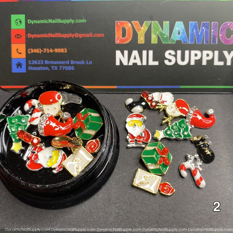 A vibrant collection of Christmas Metal Charms for Nail Art Design includes festive motifs such as Santa Claus, gifts, candy canes, and letters. Some charms are artfully arranged on a black circular surface, while others are scattered around it. In the background is a business card featuring the Dynamic Nail Supply brand and contact details.