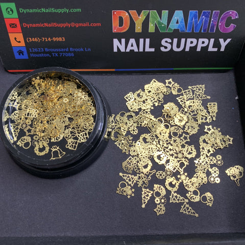 A black container holds gold Christmas Metal Sequins for Nail Art Design by Dynamic Nail Supply, featuring festive shapes such as stars, Christmas trees, snowflakes, and hearts. Some sequins are spread outside the container, adding to the holiday cheer. The backdrop highlights Dynamic Nail Supply's contact information in Houston, Texas.