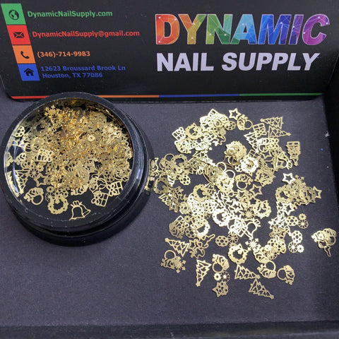 A container of the Christmas Metal Sequins for Nail Art Design from Dynamic Nail Supply overflows onto a black surface, creating festive shapes such as stars, hearts, snowflakes, and Christmas trees. In the background lies a colorful card from Dynamic Nail Supply with contact information.