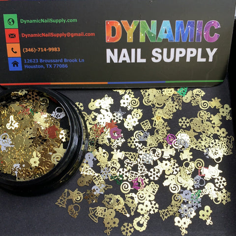 A black circular container tipped over on a table, revealing the Christmas Metal Sequins for Nail Art Designs along with various decorations like stars, hearts, and other shapes. In the background sits a business card featuring Dynamic Nail Supply's contact information and its colorful Houston, TX address.