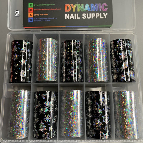 A clear plastic box containing 10 rolls of the Christmas Nail Art Foils - [02] from Dynamic Nail Supply features holographic designs with patterns such as stars, hearts, snowflakes, and festive elements on black and silver backgrounds. The lid of the box prominently displays the Dynamic Nail Supply logo and contact information.