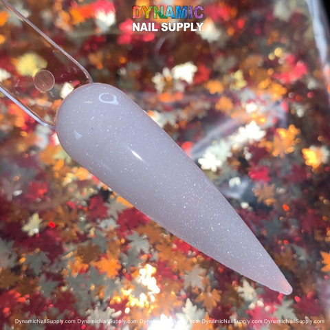 A close-up view features a sparkly, pointed nail art sample on a clear display stick, adorned in the 453 Diamond Dust acrylic powder from Dynamic Nail Supply. The nail exhibits a light pink hue with subtle glitter, set against a background of shimmering red and gold leaf patterns. Above, the name "Dynamic Nail Supply" shines brightly in colorful letters, highlighting the allure of acrylic nails.