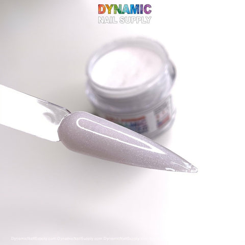 A close-up of a long, pointed nail features a light, shimmering pink polish from the 453 Diamond Dust acrylic powder collection, embodying elegant nail art. In the background, a small open jar of matching dipping powder rests on a white surface. The jar is slightly blurred to emphasize the polished acrylic nail. At the top, “Dynamic Nail Supply” is prominently displayed in colorful text.