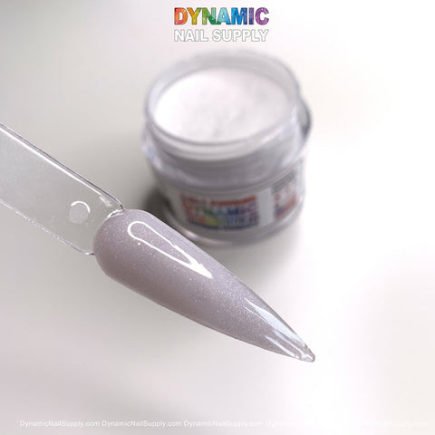 A close-up of a nail polish swatch on a translucent stick, displaying the shimmery light pink shade ideal for elegant nail art. An open jar of Dynamic Nail Supply's 453 Diamond Dust - Acrylic Powder for Dipping and Sculpting stands behind it. The background is softly illuminated, and the text "Dynamic Nail Supply" appears at the top in colorful letters.