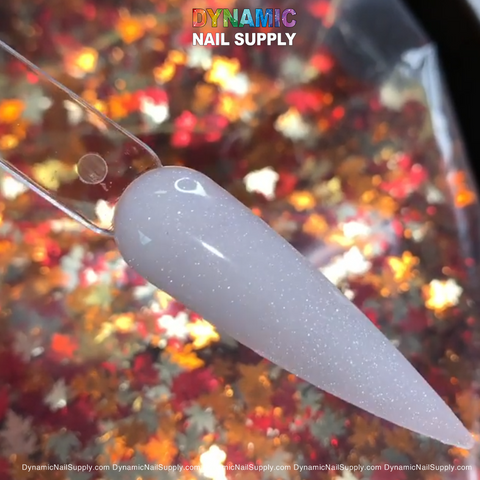 A nail tip painted with 453 Diamond Dust, a shimmery, translucent white acrylic powder from Dynamic Nail Supply, is set on a clear stick against a vibrant backdrop of red, orange, and gold maple leaf confetti. The brand name Dynamic Nail Supply is prominently featured at the top in multicolored text.