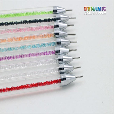 The Dual-head Rhinestone Picker Pen by Dynamic Nail Supply showcases ten dotting tools with clear handles filled with colored crystals ranging from green to red. Each tool has a silver tip and is perfect for precise nail art designs or use as a rhinestone picker. The brand name, DYNAMIC NAIL SUPPLY, is displayed in multicolored letters at the top right corner.