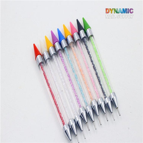 A collection of ten Dual-head Rhinestone Picker Pens by Dynamic Nail Supply is displayed, each equipped with a wax head ideal for picking up rhinestones. The pens feature clear barrels adorned with shimmering gems and vibrant caps, all organized in a gradient pattern from red to black on a white background. In the top right corner appears the brand name "Dynamic Nail Supply" in multicolored lettering.