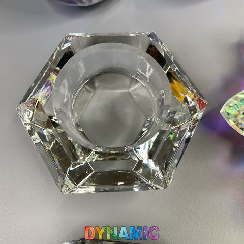 A crystal Dappen dish from Dynamic Nail Supply, beautifully designed with intricate facets in hexagon, round, heart, or lotus shapes, is centered on a gray surface. Below the dish, the brand name "Dynamic Nail Supply" is boldly displayed in colorful letters featuring a gradient from red to purple. The dish reflects light stunningly to create delicate rainbow hues.