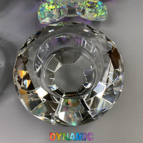 A lead crystal bowl glimmers with prismatic reflections on a gray surface. Above it, another smaller, colorful crystal object is partially visible from the CRYSTAL DAPPEN DISH collection in unique shapes like hexagon and heart. At the bottom, the word "Dynamic" appears in bold, rainbow-colored letters, representing Dynamic Nail Supply.