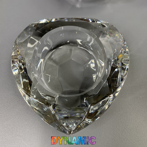 A heart-shaped clear lead crystal dish from Dynamic Nail Supply, featuring a geometric, multifaceted design reminiscent of a Dappen dish with a circular indent at its center, rests on a gray surface. Below it, the brand name "Dynamic" is displayed in colorful gradient letters.